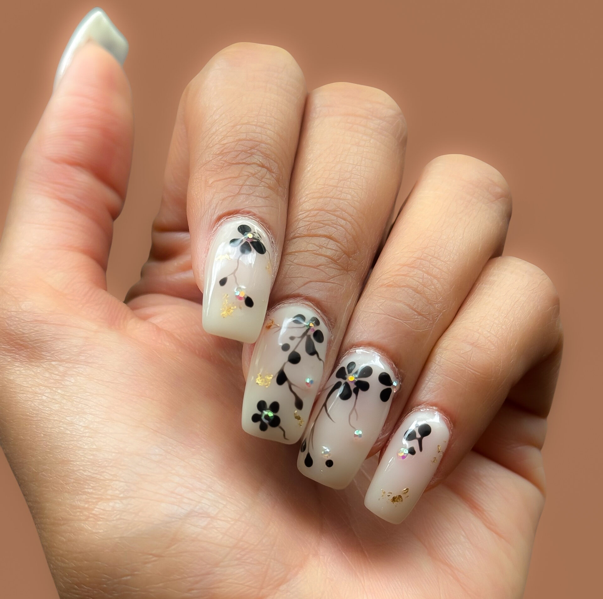 Floral gel nail art with glossy nude nails, black flower designs, gold accents, and decorative embellishments.